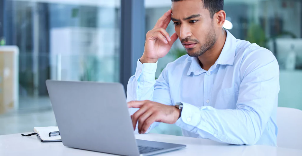 homem sentado de frente para um notebook com a mão na cabeça e expreessão de preocupaçao