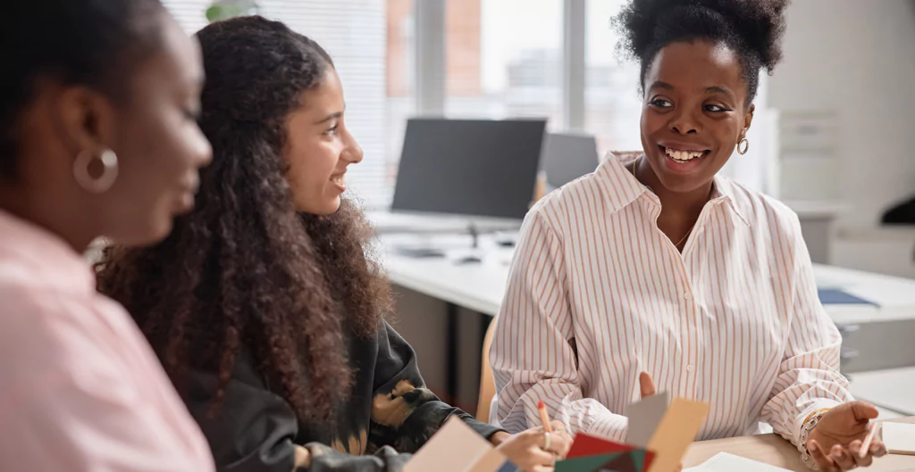 Três mulheres interagindo em um ambiente de trabalho moderno, trocando ideias e sorrisos durante uma reunião colaborativa.