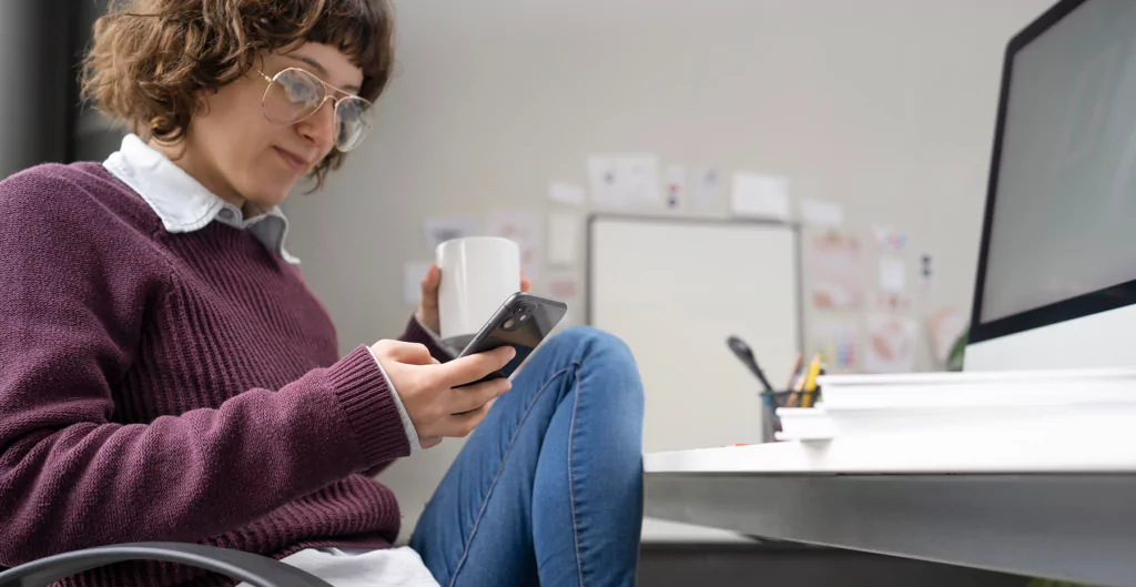 Mulher sentada em cadeira de escritório, olhando para o celular e com uma xícara de café na outra mão