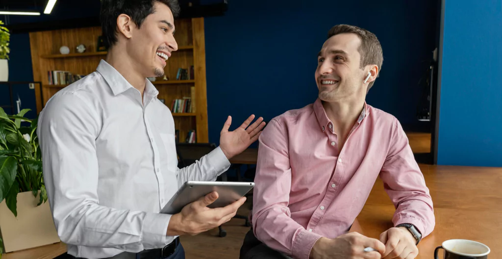 Dois homens em uma conversa alegre no ambiente de trabalho, um segurando um tablet e o outro com um sorriso, simbolizando colaboração e dinamismo.