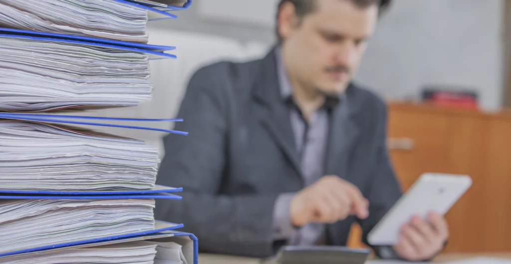 Homem segurando celular com pilha de documentos ao lado