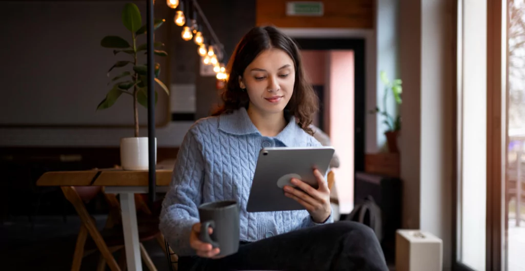 Uma jovem mulher sentada em um café, usando um suéter azul claro, segurando um copo e um tablet. O ambiente é moderno e acolhedor, com luzes pendentes ao fundo.