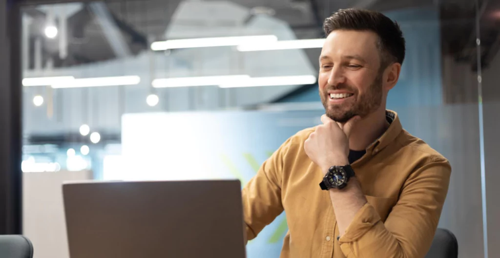 Homem sorridente trabalhando em laptop em um ambiente moderno de escritório, focado em suas atividades e com expressão de satisfação.