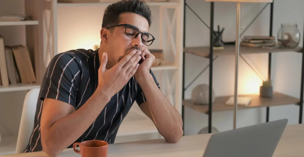 Homem com óculos, vestido com camisa listrada, aparenta estar cansado enquanto trabalha no computador. Ele se espreguiça com uma xícara na mesa.