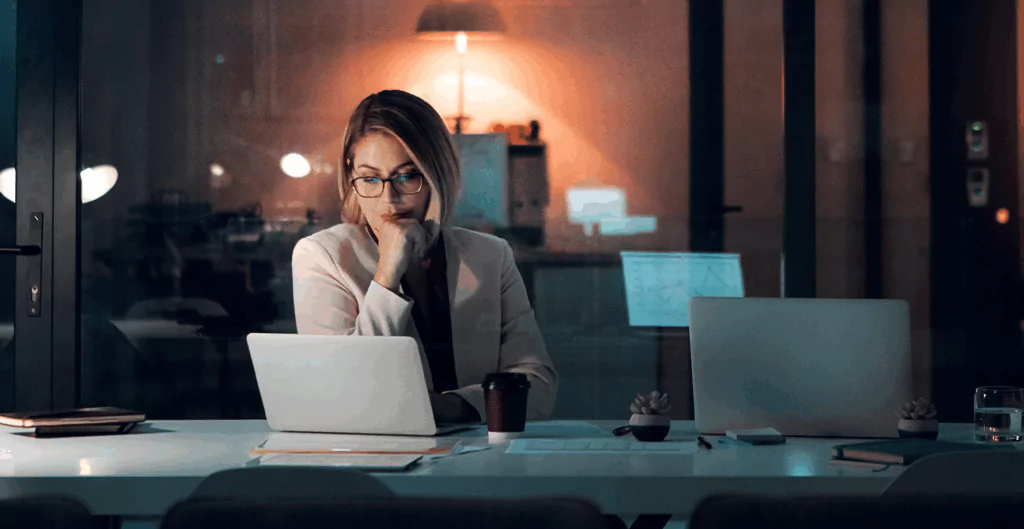 Mulher a noite, trabalhando em notebook com escritório ao fundo