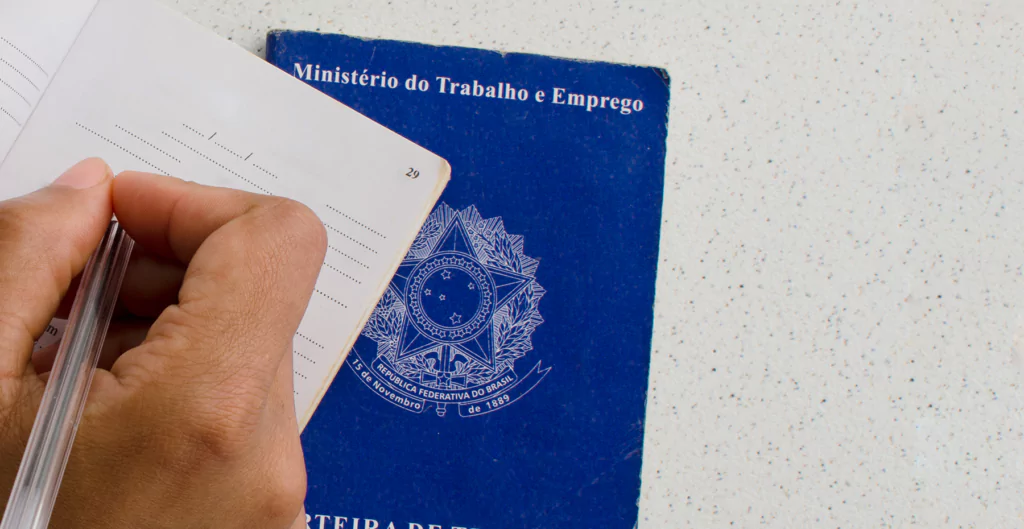 Imagem de uma mão escrevendo em uma página de um documento, com a carteira de trabalho ao fundo. A carteira é azul e possui o símbolo do Ministério do Trabalho e Emprego.
