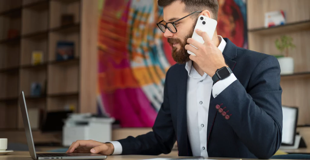 Homem de cabelos curtos e barba usando smartphone enquanto trabalha em laptop em ambiente moderno. Estilo profissional e produtivo.