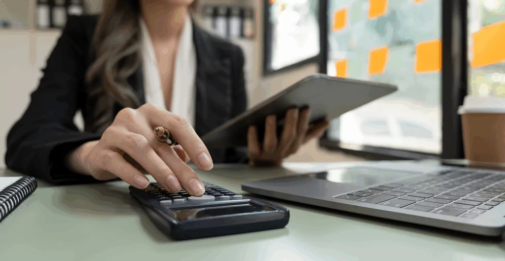 Pessoa de negócios usando calculadora e tablet em ambiente de escritório, com laptop e bloco de anotações ao lado. Imagem relacionada a planejamento financeiro, finanças pessoais ou negócios.