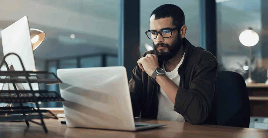 Homem pensativo usando laptop em ambiente de escritório moderno, com foco na concentração e trabalho remoto.