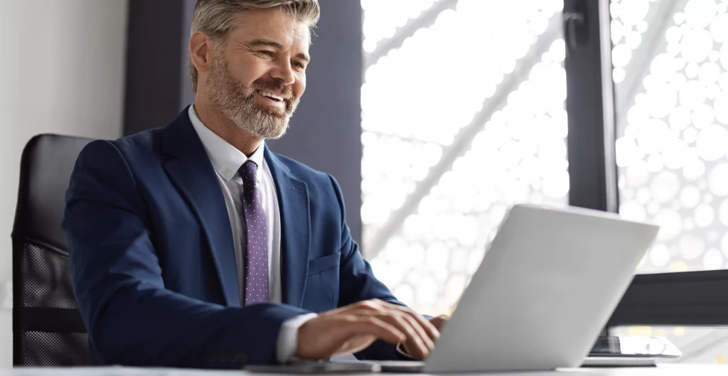 Executivo sorridente usando um laptop em um escritório moderno com iluminação natural, representando profissionalismo e tecnologia.