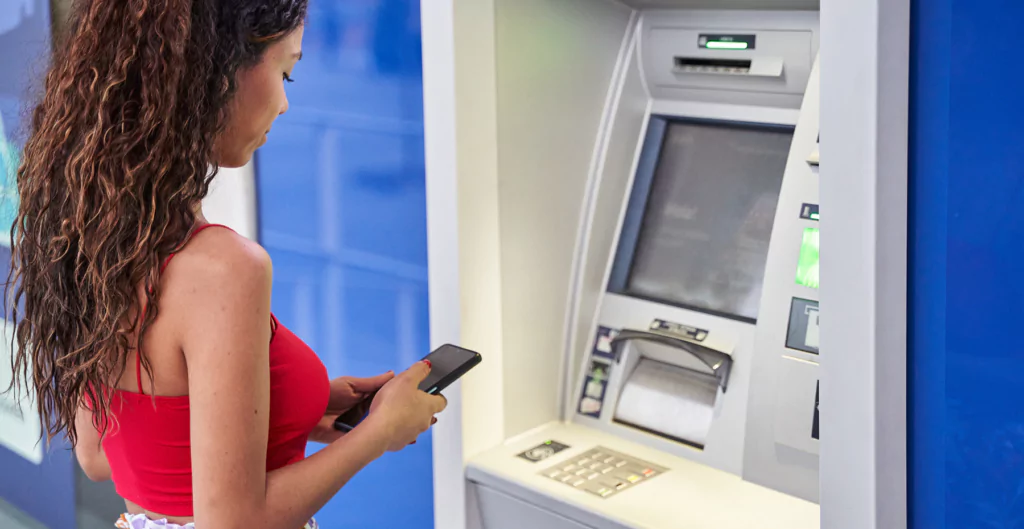 Mulher usando o celular próxima a um caixa eletrônico no banco, realizando transação bancária ou saque automático.
