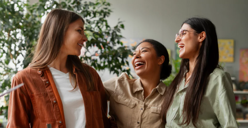 Três mulheres felizes e sorridentes interagindo em um ambiente ao ar livre, representando amizade e bem-estar.