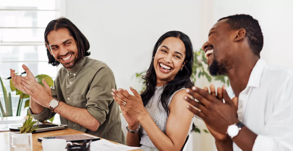 Três colegas de trabalho sorrindo e aplaudindo durante uma reunião, demonstrando felicidade e colaboração no ambiente de escritório.