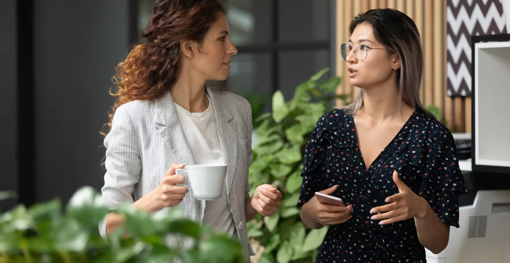 Duas mulheres conversando em um ambiente moderno, com plantas ao fundo. Uma delas segura uma xícara de café e a outra usa óculos.