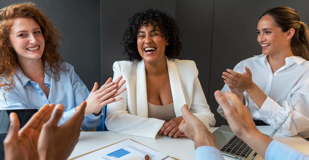 Grupo de pessoas em ambiente corporativo celebrando uma conquista. Mulheres sorrindo e aplaudindo durante reunião de negócios, transmitindo alegria e cooperação.
