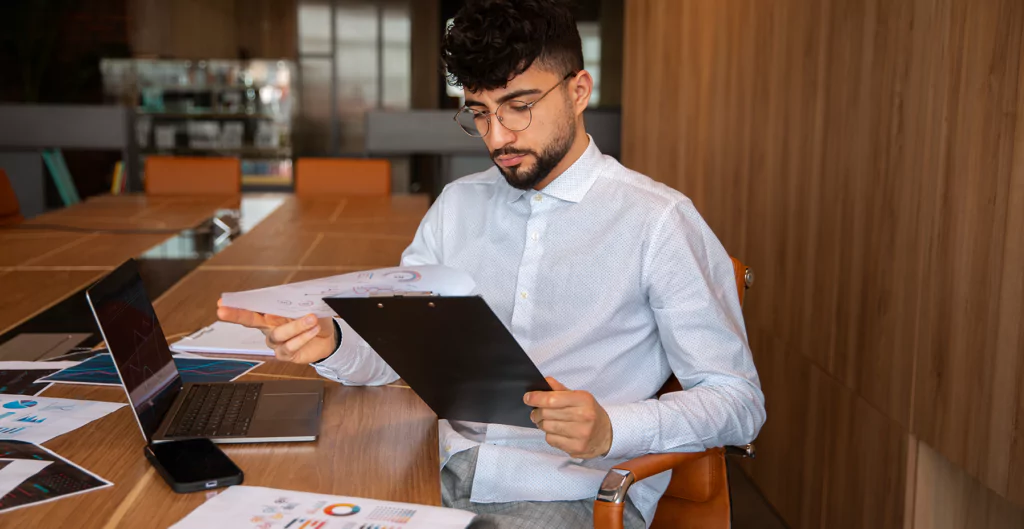 Homem de camisa azul claro analisando documentos em mesa de trabalho, laptop aberto e gráficos ao redor, representando o ambiente de negócios e produtividade.
