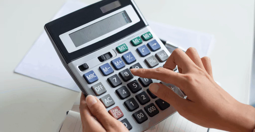 Pessoa utilizando uma calculadora de mesa para realizar cálculos financeiros ou contábeis, com uma caneta ao lado e papéis sobre a mesa.