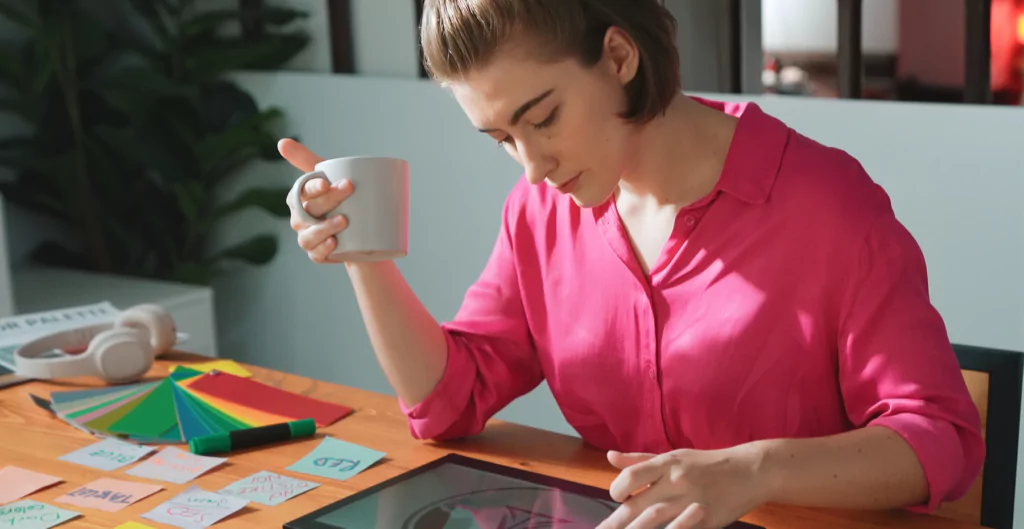 Mulher de camisa rosa, segurando uma caneca, analisando anotações e um tablet sobre uma mesa de madeira