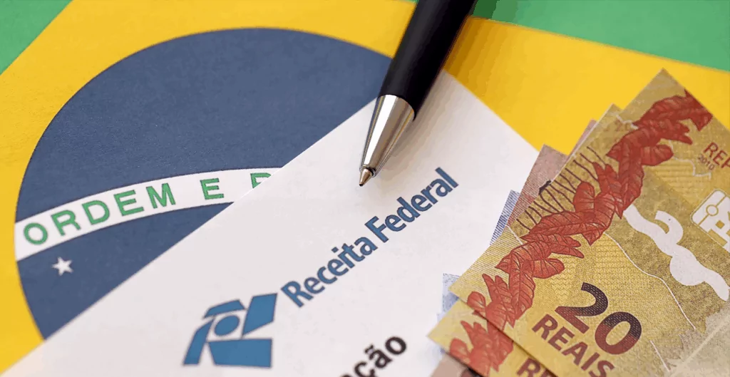 bandeira do Brasil com notas de vinte reais, uma caneta e um documento da Receita Federal 