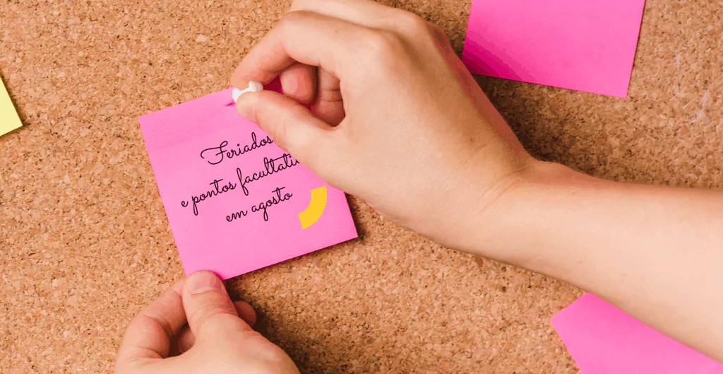 pessoa colocando post-it rosa, em quadro de avisos com a frase "feriados e pontos facultativos em agosto"