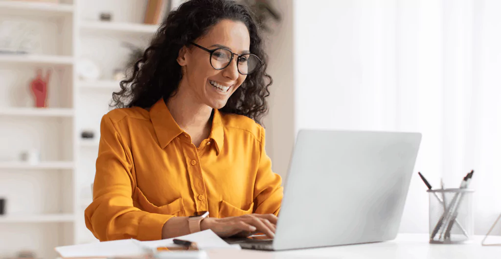 Mulher sorridente com óculos de grau trabalha no laptop em ambiente de escritório bem iluminado, demonstrando satisfação e produtividade.