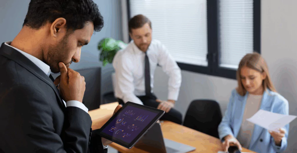 Reunião de negócios com três profissionais em um escritório, um deles analisando dados em um tablet, enquanto os outros dois observam com atenção.