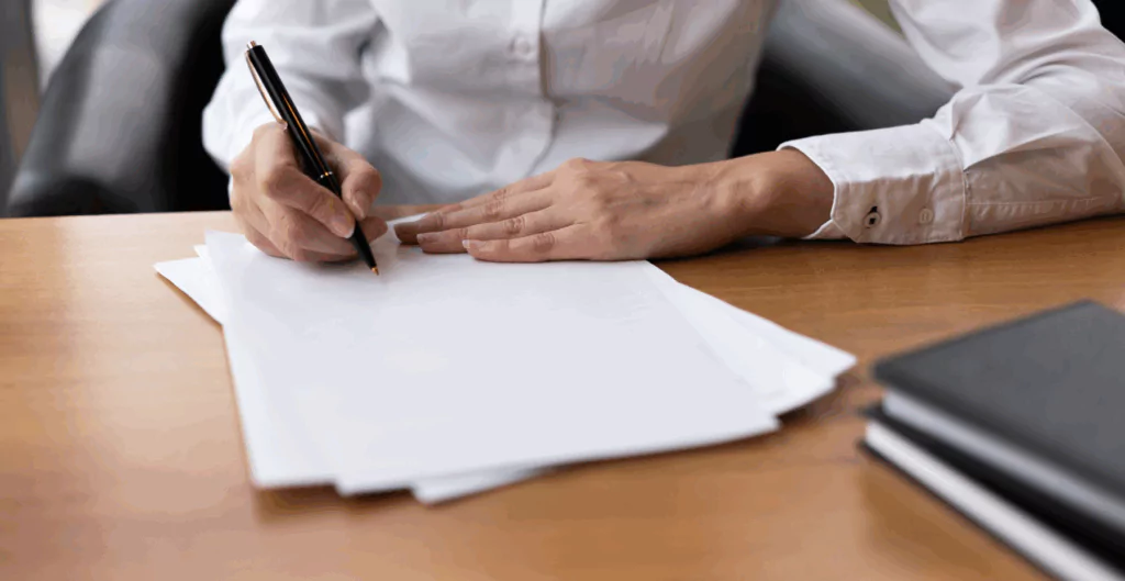 Pessoa, com roupa social, assinando uma cara de demissão sobre uma mesa de madeira 