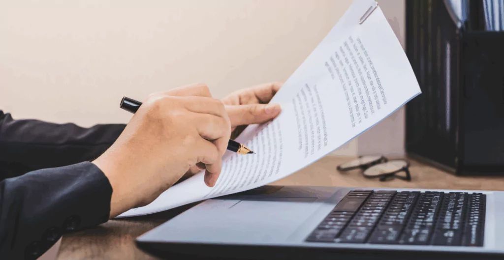 Uma mão segurando uma caneta preta e a outra uma carta de demissão com um notebook ao lado