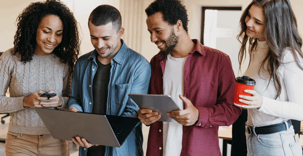 Grupo de jovens sorrindo e interagindo com dispositivos digitais em ambiente moderno, promovendo tecnologia e conexão social.