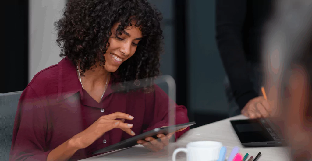 Mulher sorridente usando um tablet em uma reunião de trabalho, com ambiente profissional ao fundo.