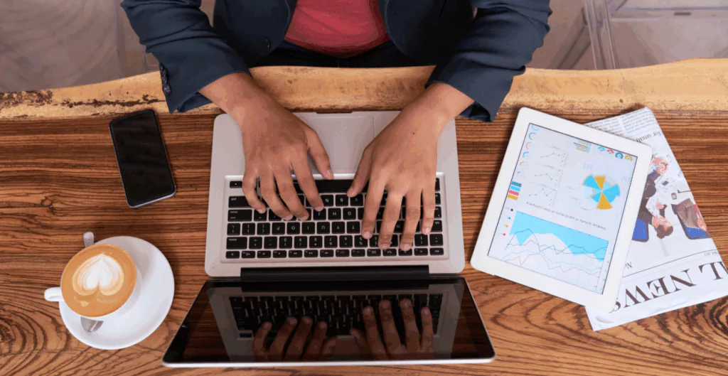 Pessoa trabalhando em um laptop com gráficos e tablet ao lado, em ambiente de escritório.