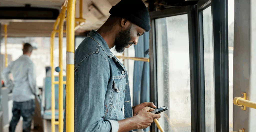 Homem usando smartphone dentro de um ônibus, trajado de jaqueta jeans e boné.