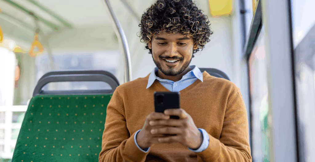 Homem sorridente usando celular em transporte público, vestindo suéter marrom e camisa azul, com cabelo cacheado e barba. 