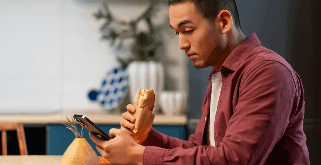 Homem comendo enquanto usa o celular, um hábito que pode contribuir para o brain rot pela falta de foco e excesso de estímulos.