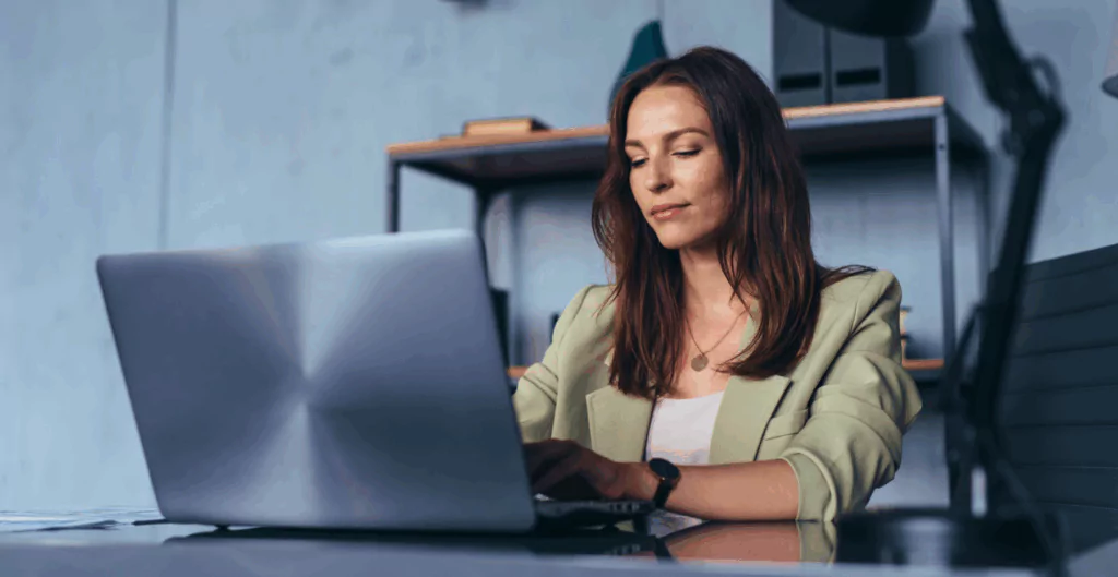 Mulher digitando em notebook com escritório ao fundo