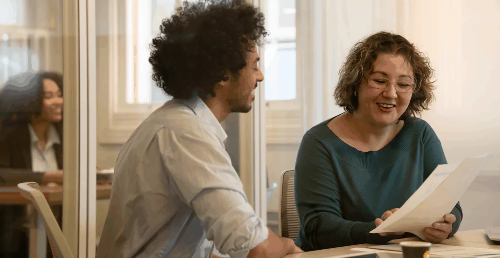 Dois colegas conversando e analisando documentos em ambiente corporativo, representando a psicodinâmica do trabalho aplicada às relações profissionais, à escuta ativa e ao fortalecimento da cooperação.