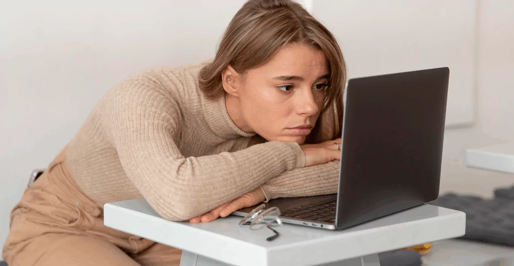 Mulher jovem apoiada sobre o notebook, com expressão de cansaço e desmotivação, ilustrando os desafios emocionais e os impactos da psicodinâmica do trabalho sobre a saúde mental no ambiente profissional.