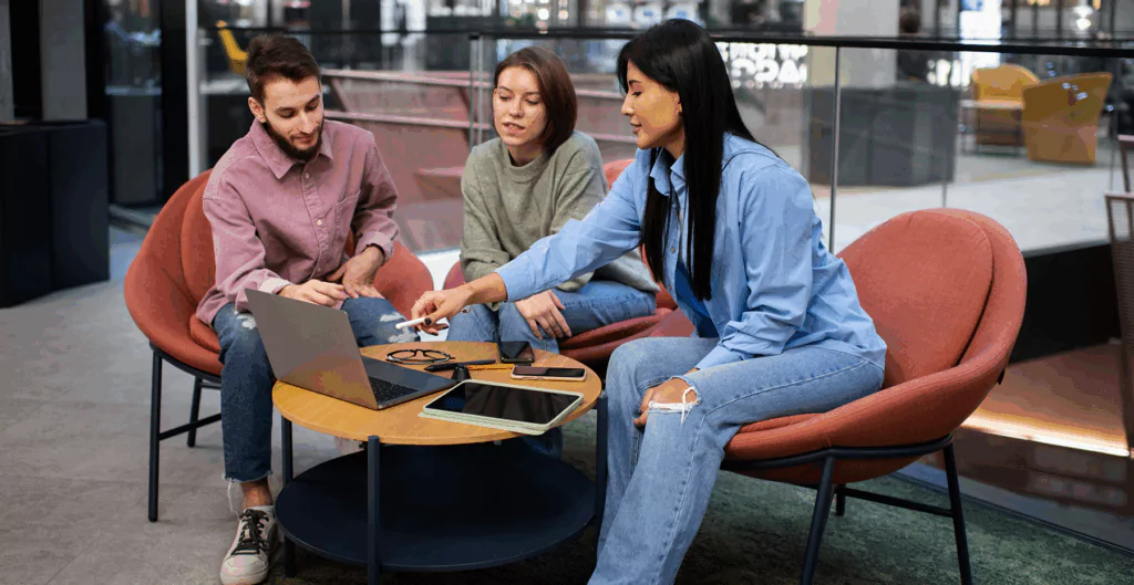 Grupo de jovens profissionais em reunião de trabalho em ambiente moderno, utilizando laptop e tablets na mesa.