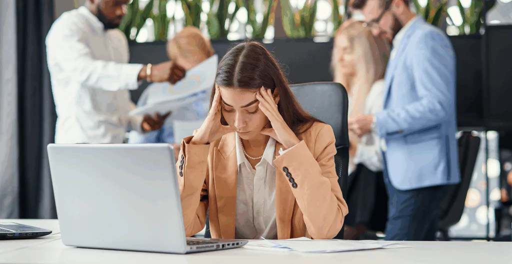 Mulher com roupas sociais, com as mãos na cabeça na frente do notebook. E pessoas conversando ao fundo em ambiente corporativo.