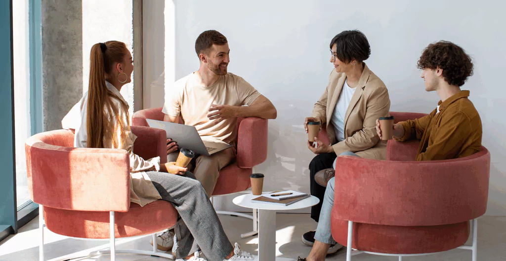 Grupo de quatro pessoas conversando e sorrindo em reunião casual com café, sentadas em cadeiras confortáveis de veludo rosa, na decoração moderna de escritório com luz natural.