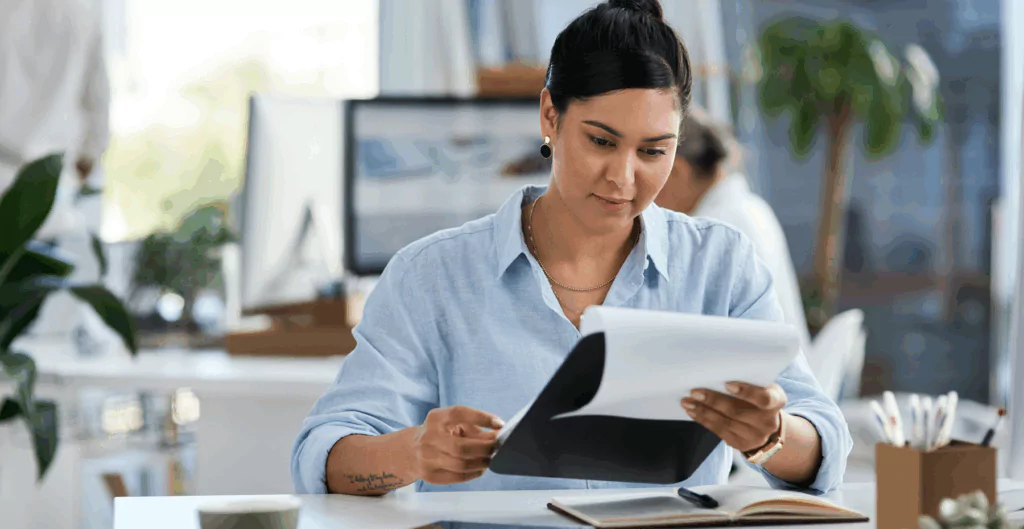 Mulher estudando documentos em escritório moderno, concentrada na análise de papéis e utilizando computador ao fundo, ambiente de trabalho organizado e produtivo.
