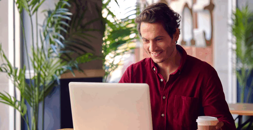 Homem sorridente usando um notebook portátil em um ambiente bem iluminado, com plantas ao fundo,