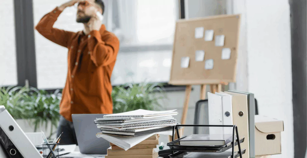 Homem de negócios estressado em escritório com pilhas de papéis, notebooks e uma lousa com post-its ao fundo, representando sobrecarga de trabalho e estresse profissional.