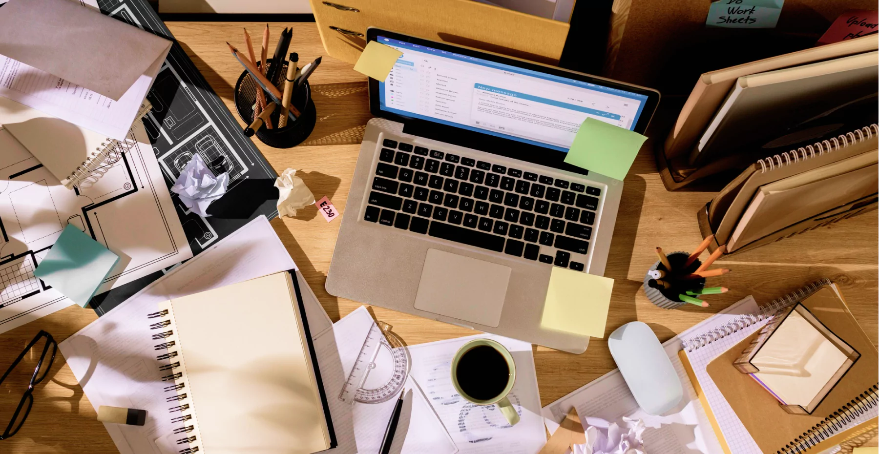 Mesa de trabalho estudantil ou de escritório com laptop, caderno, canetas, papéis, xícara de café e materiais de oficina, tudo organizado de forma caótica.