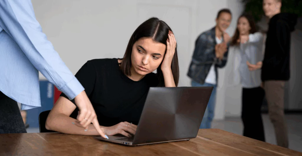 Pessoa apontando para notebook de uma outra mulher com rosto frustrado. Com outras pessoas rindo ao fundo