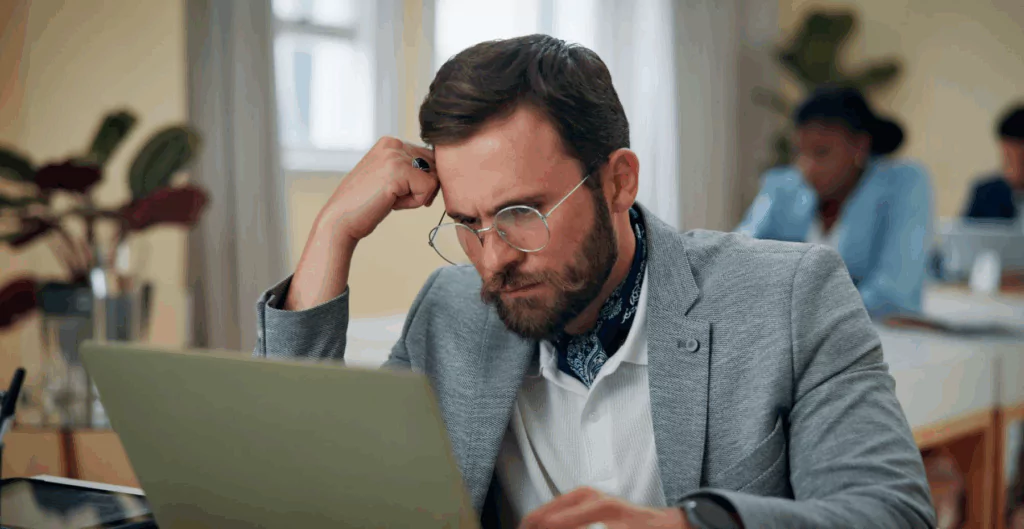 Homem com rosto preocupado para notebook, com escritório corporativo ao fundo