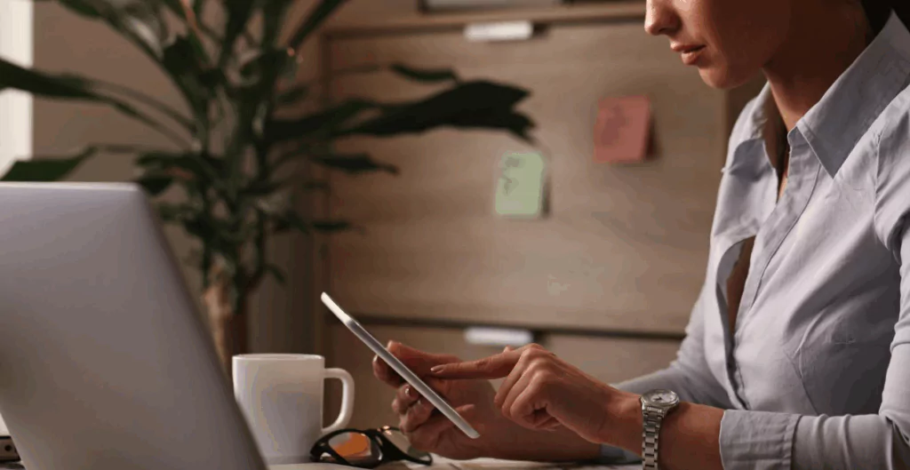 Mulher de negócios usando smartphone em escritório moderno, com laptops e caneca de café, ambiente profissional e organizado.