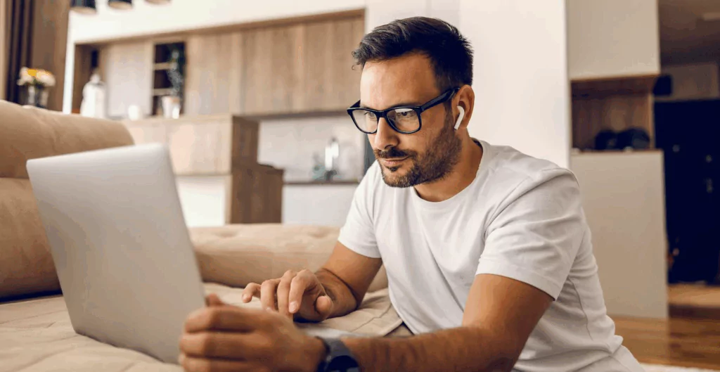 	
Homem usando óculos e fones de ouvido, trabalhando ou assistindo conteúdo no laptop em casa, ambiente moderno e aconchegante.