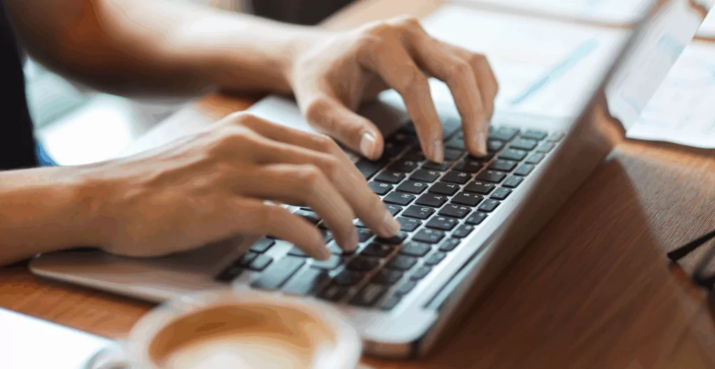 Mãos digitando em um notebook sobre uma mesa com xícara de café ao lado