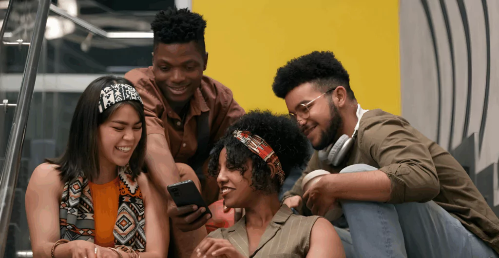 Quatro jovens sorrindo para um celular, sentados em uma escada